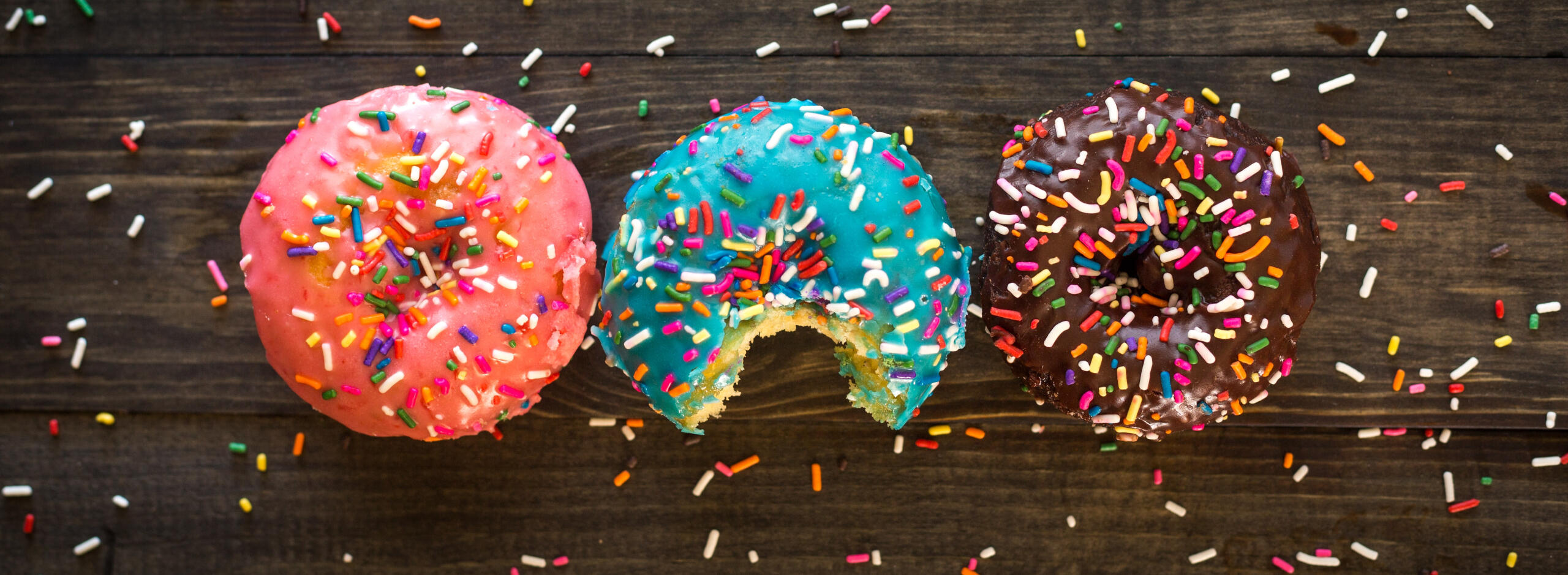 3 colourful iced doughnuts with sprinkles on them and on the wooden worktop, viewed from above. Photo by Patrick Fore on Unsplash